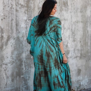 Pode incluir: Um vestido maxi azul turquesa e marrom com estampa tie-dye e uma silhueta longa e fluida. O vestido tem um decote em V e mangas curtas. A modelo est&aacute; vestindo o vestido e est&aacute; em p&eacute; em frente a uma parede de concreto. O texto "KAFTANBUZZ" &eacute; vis&iacute;vel no vestido.