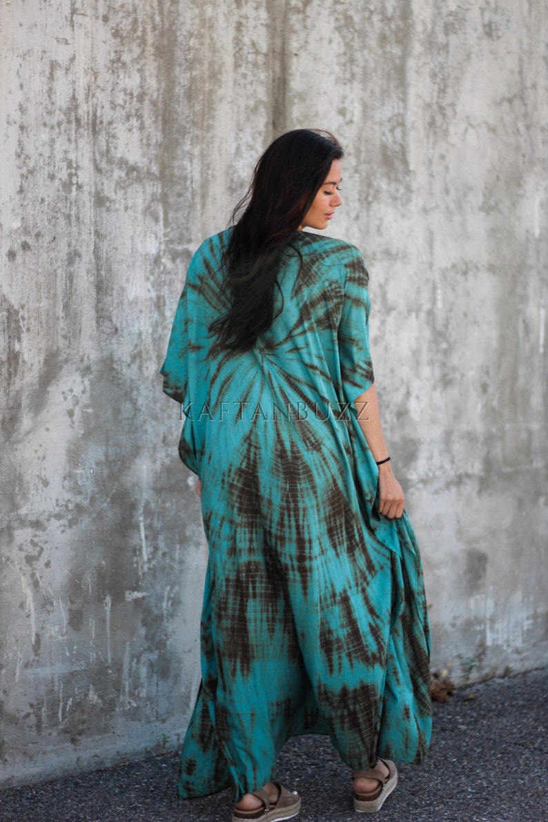 Pode incluir: Um vestido maxi azul turquesa e marrom com estampa tie-dye e uma silhueta longa e fluida. O vestido tem um decote em V e mangas curtas. A modelo est&aacute; vestindo o vestido e est&aacute; em p&eacute; em frente a uma parede de concreto. O texto "KAFTANBUZZ" &eacute; vis&iacute;vel no vestido.