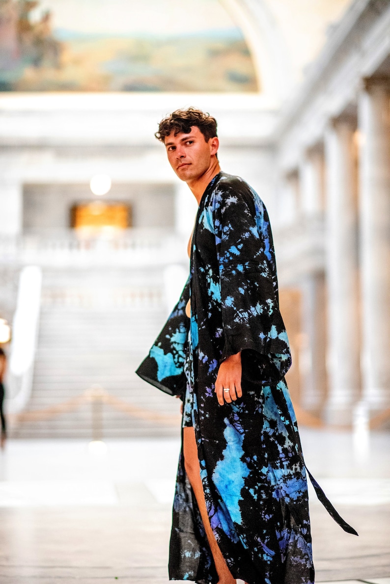 May include: A person wearing a blue and black tie-dye robe with a long, flowing design. The robe is open in the front, revealing the person's chest and arms. The person is standing in a large, white room with a high ceiling.