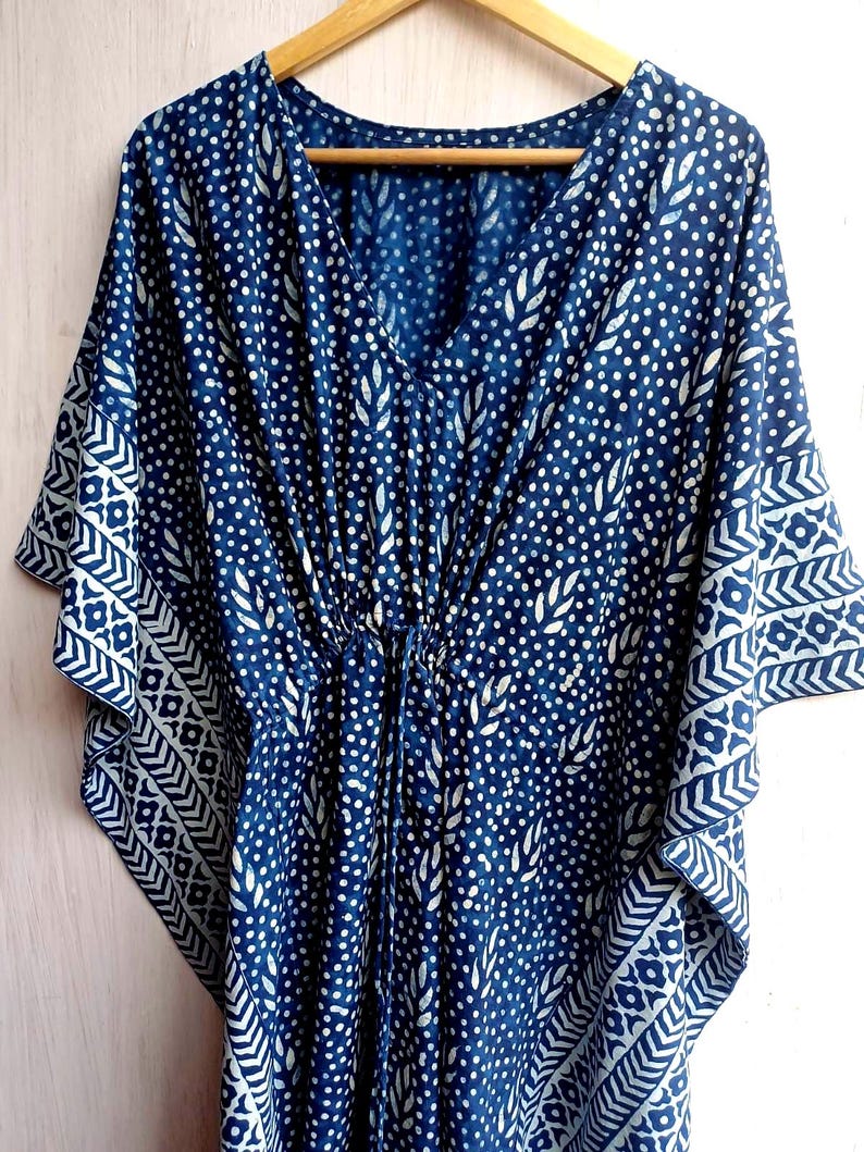 Op de afbeelding: Een blauw en wit patroon kaftan jurk met een V-hals en een taille met trekkoord. De stof heeft een herhalend patroon van witte stippen en bladvormen, met een geometrisch randontwerp op de mouwen.