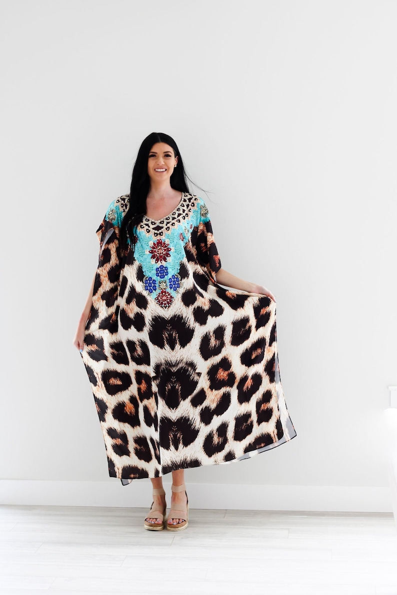 K&ouml;nnte beinhalten: Ein langes, flie&szlig;endes Maxikleid mit Leopardenmuster. Das Kleid hat ein t&uuml;rkisfarbenes und blaues Blumendesign mit Strasssteinen am Ausschnitt. Das Kleid wird von einer Person mit langen schwarzen Haaren getragen und steht auf einem wei&szlig;en Boden.