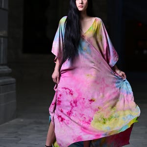 Może przedstawiać: Długa, zwiewna sukienka z żywym wzorem tie-dye w odcieniach r&oacute;żu, ż&oacute;łci i błękitu. Sukienka ma dekolt w serek i szerokie rękawy. Modelka ma na sobie czarne sandały na wysokim obcasie.