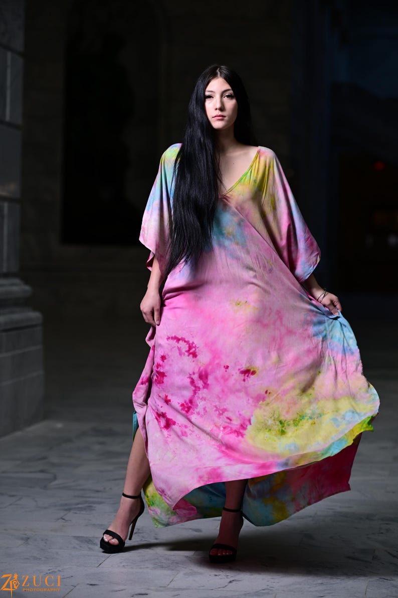 Może przedstawiać: Długa, zwiewna sukienka z żywym wzorem tie-dye w odcieniach r&oacute;żu, ż&oacute;łci i błękitu. Sukienka ma dekolt w serek i szerokie rękawy. Modelka ma na sobie czarne sandały na wysokim obcasie.