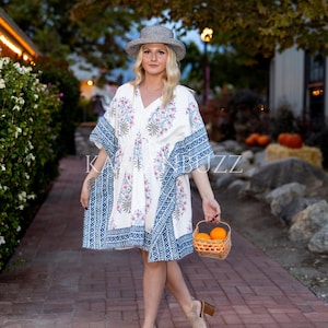 Puede incluir: Un vestido caftán blanco hasta la rodilla con estampados florales azules y rosas y bordes geométricos azules. El vestido se lleva con un sombrero de paja y tacones beige. Se sostiene una pequeña cesta de naranjas en la mano.