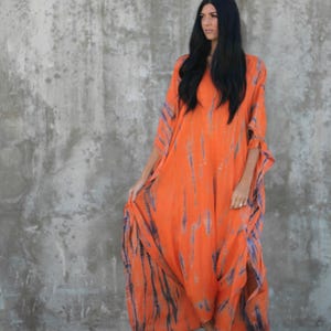 May include: A woman wearing a long, orange and blue tie-dye maxi dress with a loose, flowing silhouette. She is standing in front of a concrete wall and wearing sandals.
