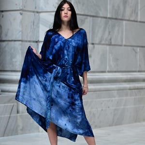 May include: A blue tie-dye dress with a V-neck and a matching belt. The dress has short sleeves and a flowing skirt. The model is wearing black high-heeled sandals. The background is a light gray marble wall.