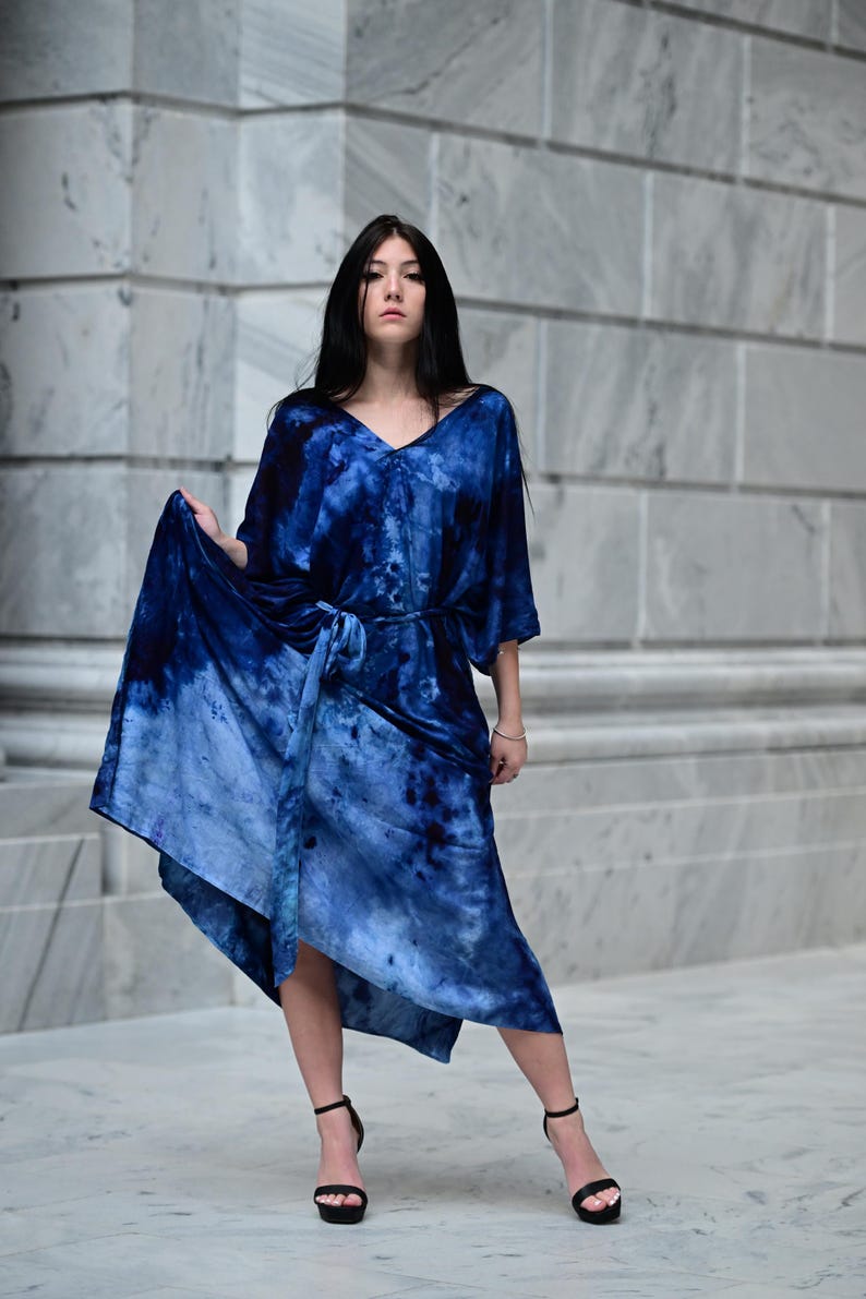 May include: A blue tie-dye dress with a V-neck and a matching belt. The dress has short sleeves and a flowing skirt. The model is wearing black high-heeled sandals. The background is a light gray marble wall.