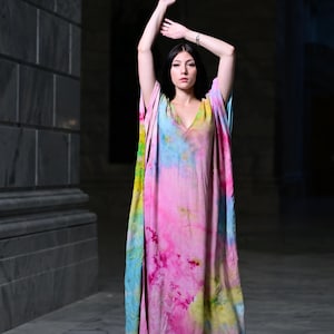 May include: A long, flowing tie-dye dress in shades of pink, blue, yellow, and green. The dress has a V-neck and wide sleeves. The model is standing with arms raised, showcasing the dress's vibrant colors and flowing fabric.