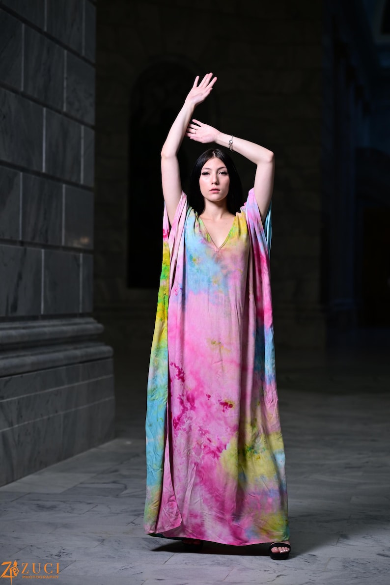 Może przedstawiać: Długa, zwiewna sukienka w stylu tie-dye w odcieniach r&oacute;żu, błękitu, ż&oacute;łci i zieleni. Sukienka ma dekolt w serek i szerokie rękawy. Modelka stoi z podniesionymi ramionami, prezentując żywe kolory i zwiewny materiał sukienki.