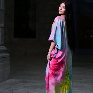 Może przedstawiać: Długa, zwiewna sukienka z żywym wzorem tie-dye w odcieniach r&oacute;żu, błękitu i ż&oacute;łci. Sukienka ma luźny, wygodny kr&oacute;j i kr&oacute;tkie rękawy. Modelka ma na sobie sukienkę w plenerze.