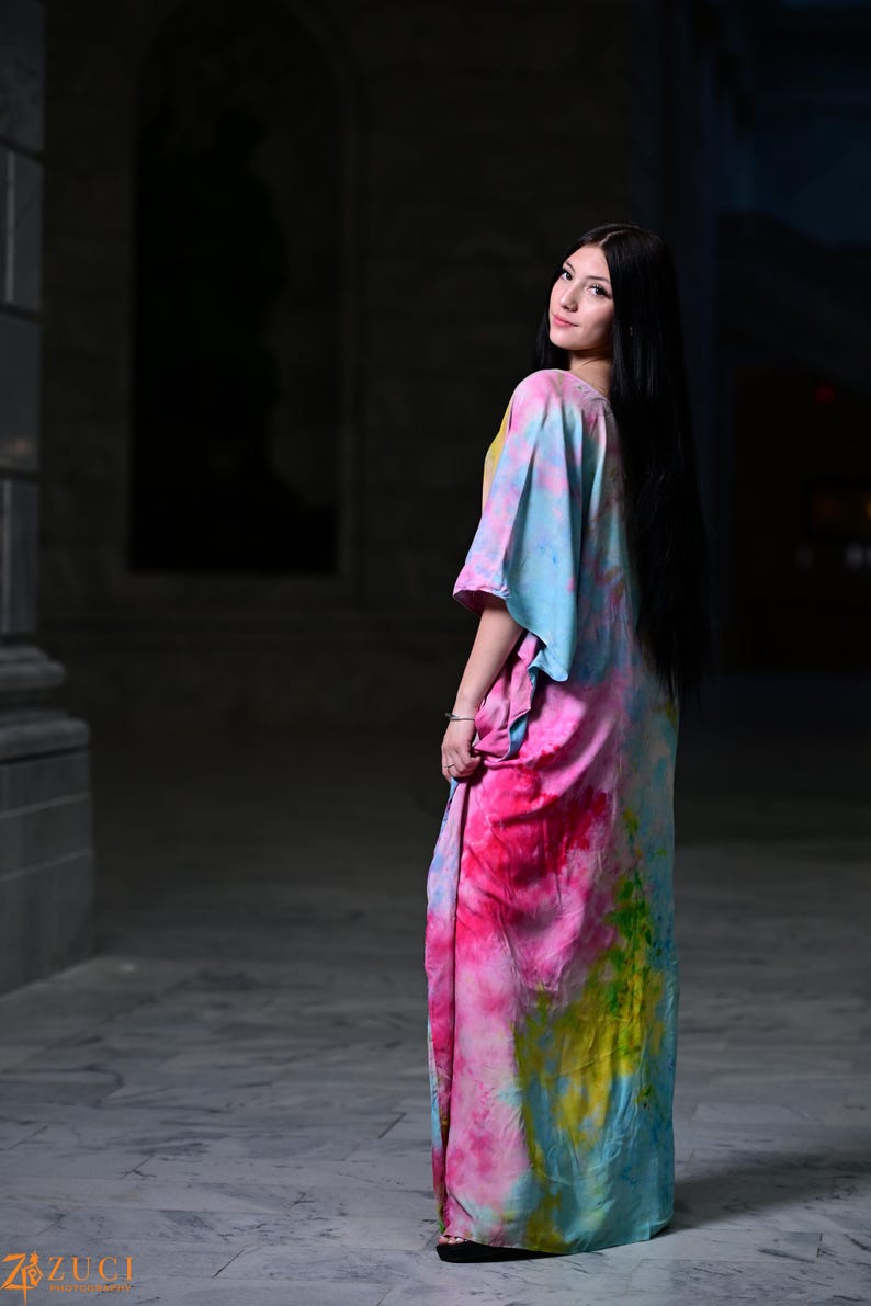 Może przedstawiać: Długa, zwiewna sukienka z żywym wzorem tie-dye w odcieniach r&oacute;żu, błękitu i ż&oacute;łci. Sukienka ma luźny, wygodny kr&oacute;j i kr&oacute;tkie rękawy. Modelka ma na sobie sukienkę w plenerze.