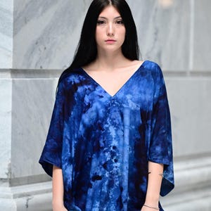 May include: A blue tie-dye V-neck tunic. The tunic has short sleeves and a loose fit. The fabric features a swirling pattern of dark and light blue hues. The model is standing in front of a marble wall.