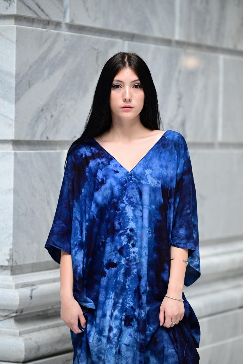 May include: A blue tie-dye V-neck tunic. The tunic has short sleeves and a loose fit. The fabric features a swirling pattern of dark and light blue hues. The model is standing in front of a marble wall.