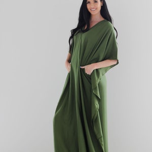 May include: A woman wearing a long, olive green, loose-fitting maxi dress with a V-neck and short sleeves. She is standing on a light wood floor and wearing tan sandals.
