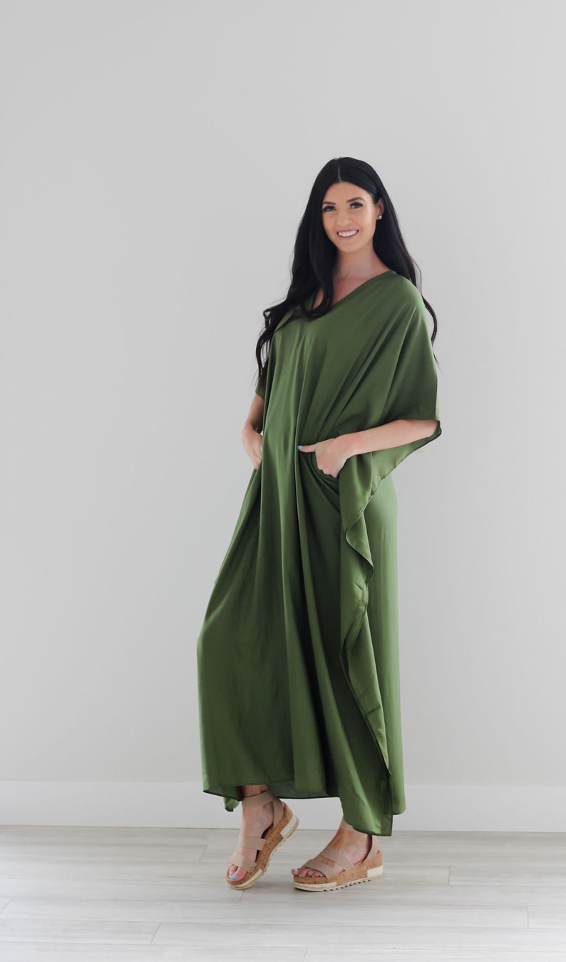 May include: A woman wearing a long, olive green, loose-fitting maxi dress with a V-neck and short sleeves. She is standing on a light wood floor and wearing tan sandals.