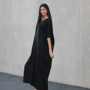 May include: A woman wearing a long, black, flowing dress with a v-neck and short sleeves. She is standing in front of a gray wall and wearing sandals.