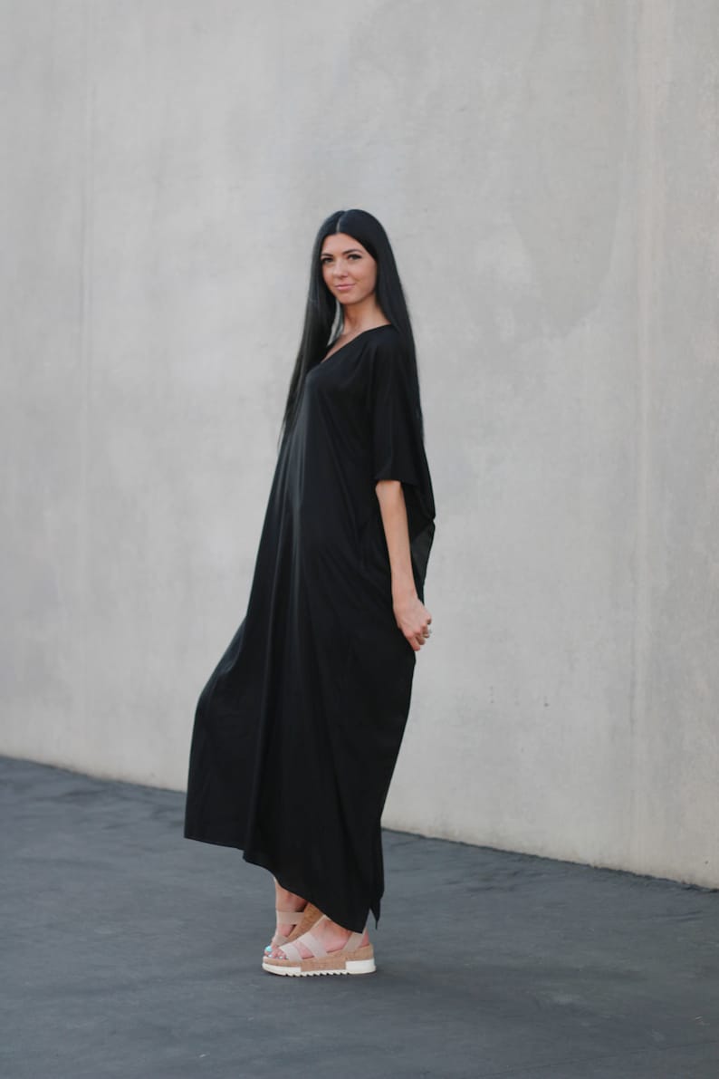 May include: A woman wearing a long, black, flowing dress with a v-neck and short sleeves. She is standing in front of a gray wall and wearing sandals.
