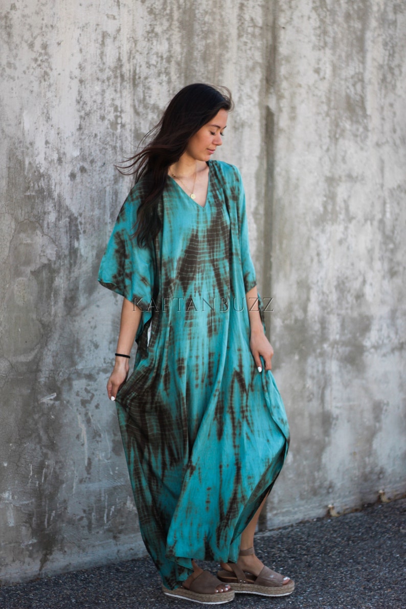 Pode incluir: Um vestido kaftan turquesa e marrom com estampa tie-dye e decote em V. O vestido tem uma silhueta fluida e &eacute; usado por uma pessoa com cabelo comprido e escuro. O vestido &eacute; fotografado contra uma parede de concreto.