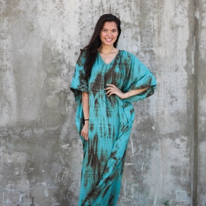 Pode incluir: Uma mulher vestindo uma longa kaftan turquesa e marrom com estampa tie-dye e decote em V. Ela est&aacute; em p&eacute; em frente a uma parede de concreto e est&aacute; usando sand&aacute;lias marrons.