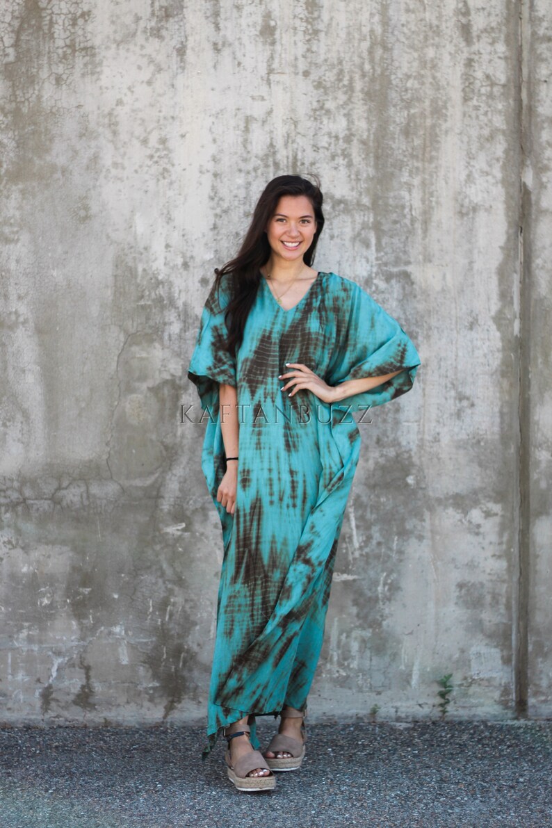 Pode incluir: Uma mulher vestindo uma longa kaftan turquesa e marrom com estampa tie-dye e decote em V. Ela est&aacute; em p&eacute; em frente a uma parede de concreto e est&aacute; usando sand&aacute;lias marrons.