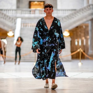 May include: A person wearing a long, black and blue tie-dye robe with a V-neck and a sash. The robe has a side slit and the person is wearing white shoes.