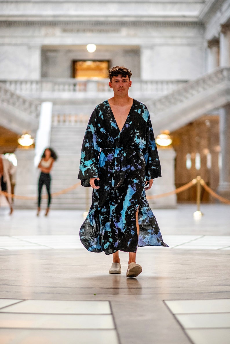 May include: A person wearing a long, black and blue tie-dye robe with a V-neck and a sash. The robe has a side slit and the person is wearing white shoes.