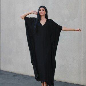May include: A woman wearing a black maxi dress with a V-neckline and long sleeves. She is standing in front of a gray wall and has her hair pulled back.