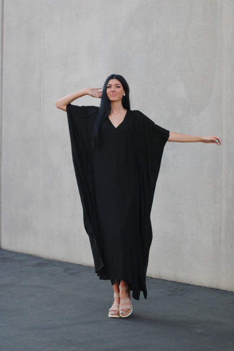 May include: A woman wearing a black maxi dress with a V-neckline and long sleeves. She is standing in front of a gray wall and has her hair pulled back.