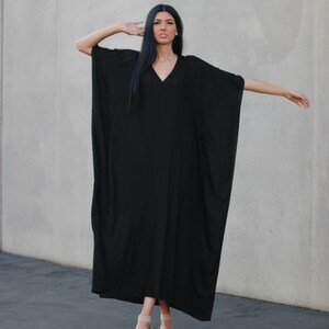 May include: A black maxi dress with a V-neckline and long sleeves. The dress is flowy and has a relaxed fit. The model is wearing the dress with nude sandals.