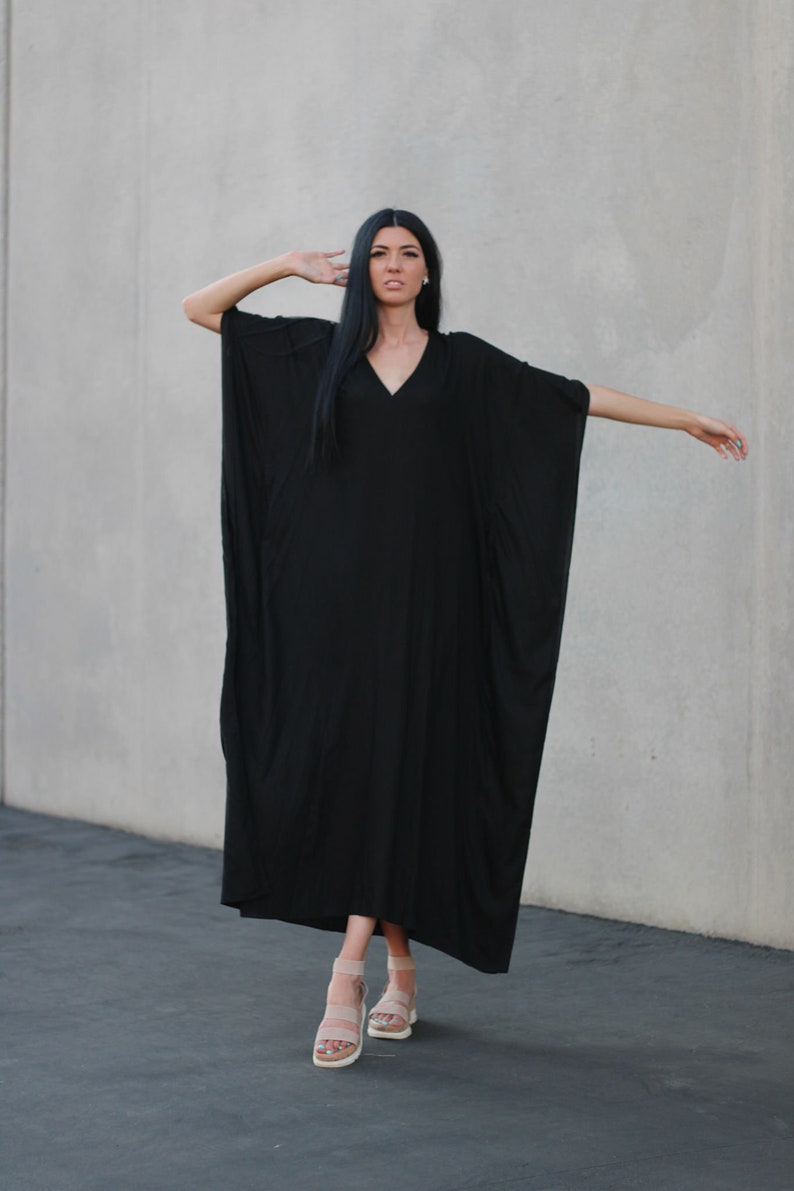 May include: A black maxi dress with a V-neckline and long sleeves. The dress is flowy and has a relaxed fit. The model is wearing the dress with nude sandals.