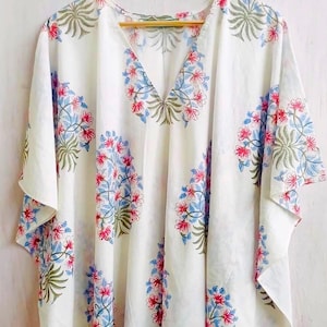 May include: White caftan with a V-neck and short sleeves, decorated with a floral pattern in red, blue, and green. The flowers and leaves are arranged symmetrically across the fabric. The garment is hanging on a wooden hanger.