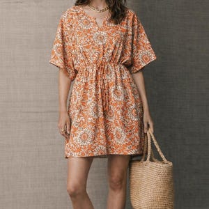 May include: Orange floral print dress with a drawstring waist and short sleeves. The dress is paired with a woven straw tote bag. The model is wearing sunglasses and a gold necklace.