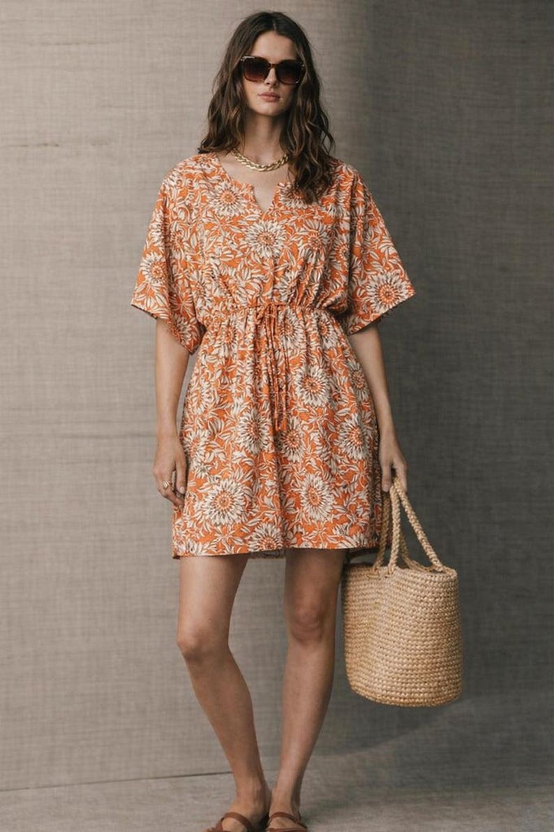 May include: Orange floral print dress with a drawstring waist and short sleeves. The dress is paired with a woven straw tote bag. The model is wearing sunglasses and a gold necklace.