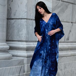 May include: A long, flowing, blue tie-dye dress with a V-neck. The dress features a gradient of blues, from deep indigo to lighter shades. The model is wearing black high-heeled sandals. The dress is made of a soft, flowing fabric.
