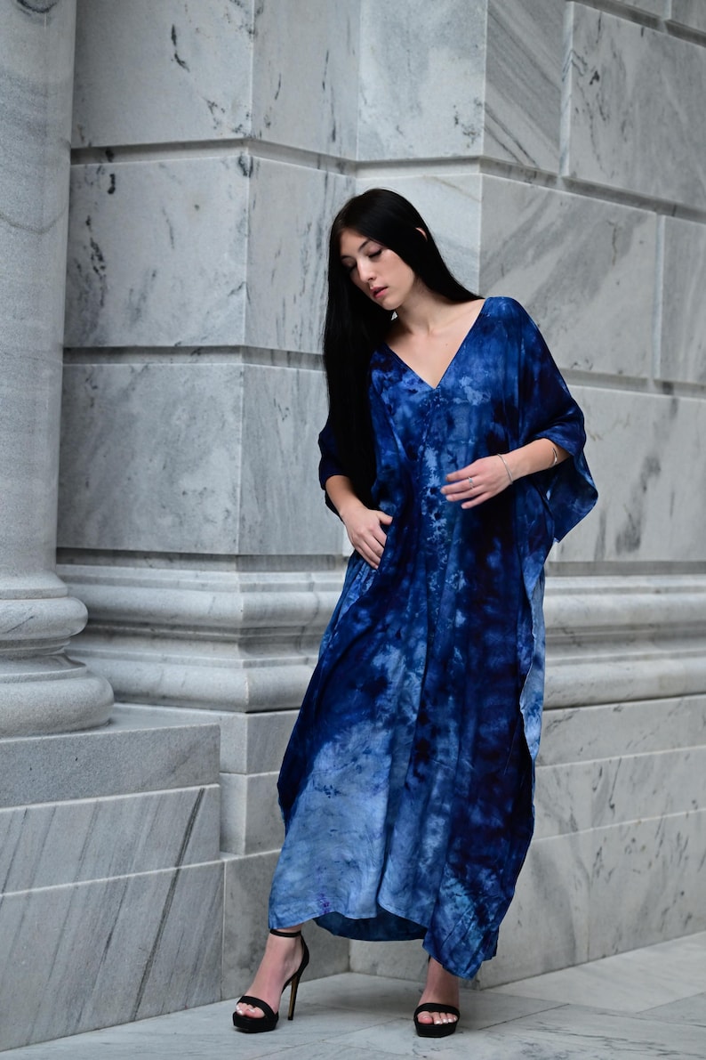 May include: A long, flowing, blue tie-dye dress with a V-neck. The dress features a gradient of blues, from deep indigo to lighter shades. The model is wearing black high-heeled sandals. The dress is made of a soft, flowing fabric.