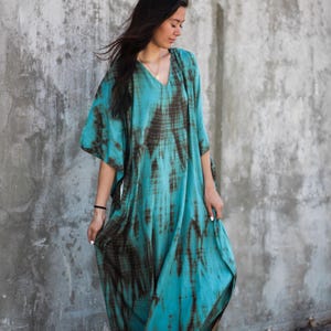 May include: A woman wearing a long, flowing turquoise and brown tie-dye dress with a V-neckline. She is standing in front of a concrete wall and is wearing brown sandals.