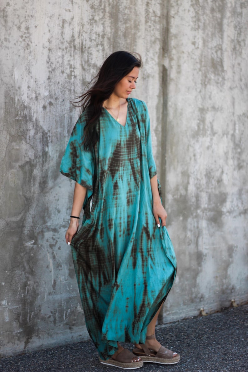 May include: A woman wearing a long, flowing turquoise and brown tie-dye dress with a V-neckline. She is standing in front of a concrete wall and is wearing brown sandals.