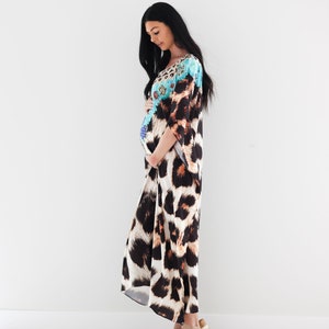 K&ouml;nnte beinhalten: Eine Frau tr&auml;gt ein langes, flie&szlig;endes Maxikleid mit Leopardenmuster. Das Kleid hat ein blaues und wei&szlig;es Blumenmuster auf der oberen H&auml;lfte. Sie steht vor einer wei&szlig;en Wand und tr&auml;gt braune Sandalen.