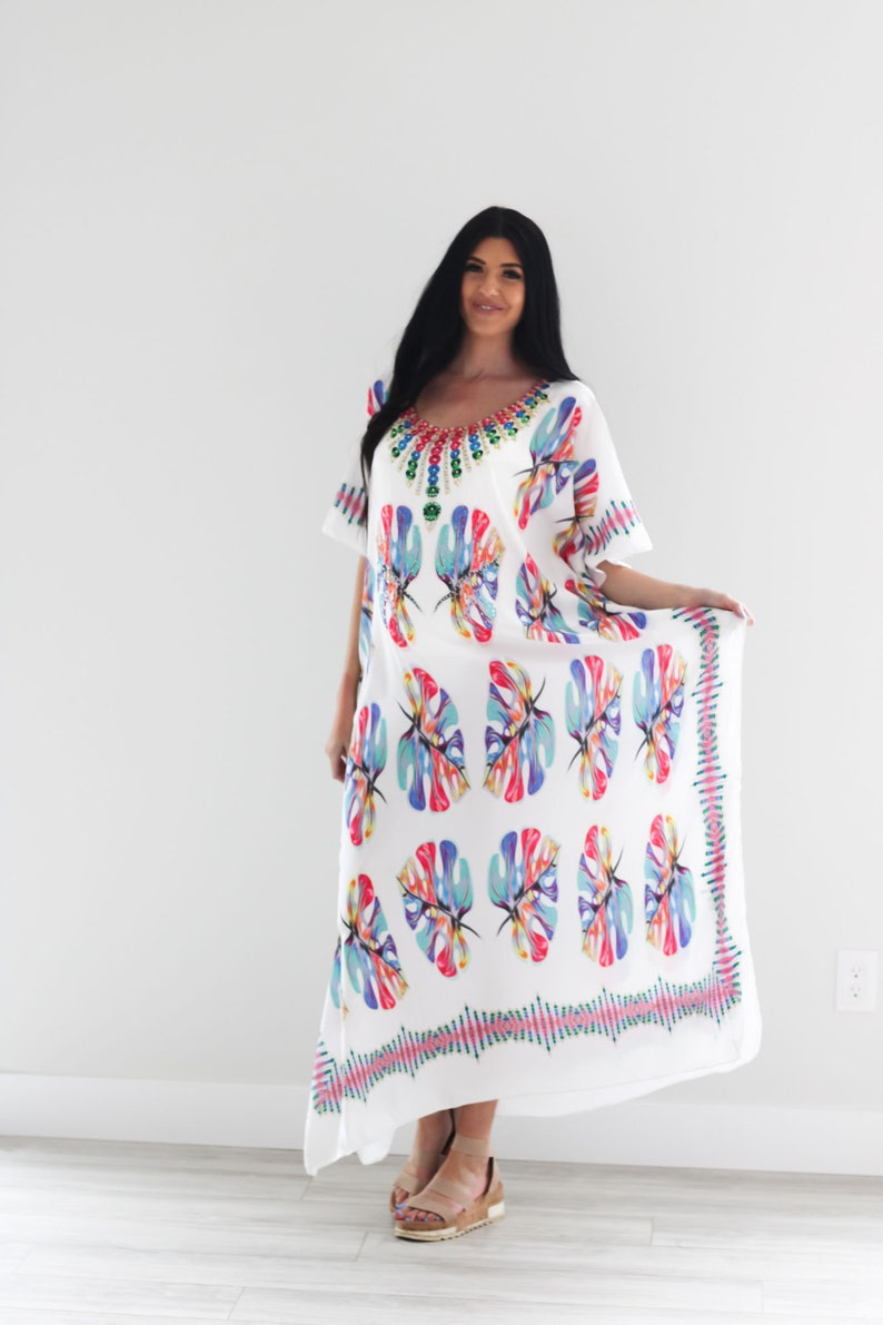 May include: White maxi dress with a colourful floral print and a beaded neckline. The dress has a flowing silhouette and a relaxed fit.