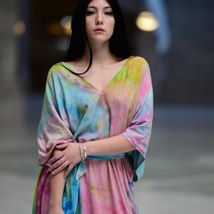 Może przedstawiać: Sukienka maxi w stylu tie-dye w odcieniach r&oacute;żu, błękitu, ż&oacute;łci i zieleni. Sukienka ma dekolt w serek, szerokie rękawy i pasujący niebieski pasek. Srebrna bransoletka zdobi nadgarstek.