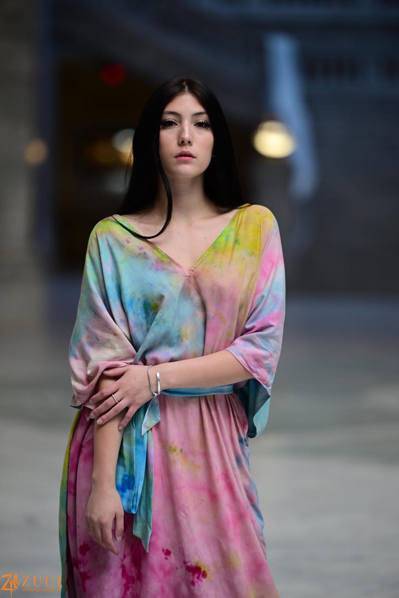 Może przedstawiać: Sukienka maxi w stylu tie-dye w odcieniach r&oacute;żu, błękitu, ż&oacute;łci i zieleni. Sukienka ma dekolt w serek, szerokie rękawy i pasujący niebieski pasek. Srebrna bransoletka zdobi nadgarstek.