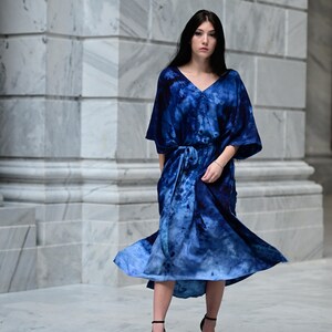 May include: A flowing, tie-dye blue dress with a V-neck and a matching belt. The dress has short sleeves and a midi length. The model is wearing black high-heeled sandals. The background is a marble wall.