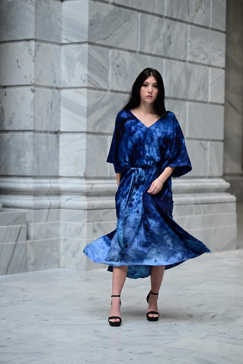 May include: A flowing, tie-dye blue dress with a V-neck and a matching belt. The dress has short sleeves and a midi length. The model is wearing black high-heeled sandals. The background is a marble wall.