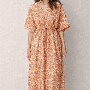 May include: An orange floral print dress with short sleeves and a V-neck. The dress has a drawstring waist and falls to the mid-calf. The model is wearing a gold necklace and brown sandals. The dress is made of a lightweight fabric.