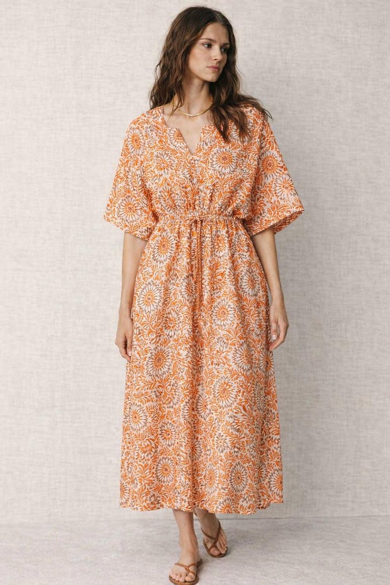 May include: An orange floral print dress with short sleeves and a V-neck. The dress has a drawstring waist and falls to the mid-calf. The model is wearing a gold necklace and brown sandals. The dress is made of a lightweight fabric.
