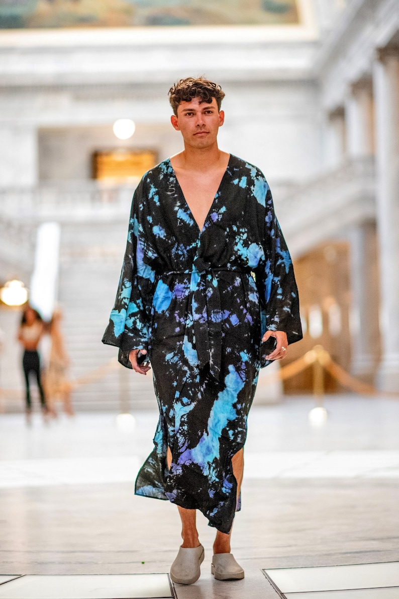 May include: A person wearing a long, black and blue tie-dye robe with a V-neck and a belt. The robe has a flowing, loose fit and is made of a soft, comfortable fabric.