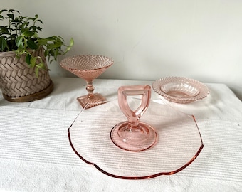 Puede incluir: Una colección de piezas de servicio de vidrio rosa, que incluyen una fuente para pasteles con asa, un cuenco de postre con pie y un cuenco pequeño con borde festoneado. Una planta en maceta está en el fondo.