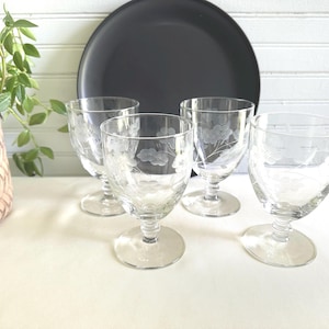 Puede incluir: Cuatro copas de cristal transparente grabadas con diseños florales están dispuestas frente a un plato negro. Las copas tienen un cuenco redondeado y un tallo corto. Una planta en maceta es visible en el lado izquierdo de la imagen.