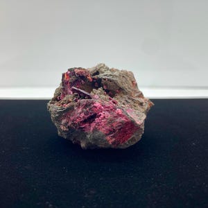 May include: A rough, gray rock formation with vibrant, hot pink crystalline structures. The mineral specimen has a rugged texture and a striking contrast between the gray matrix and the bright pink mineral deposits.