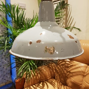 May include: A vintage industrial style pendant light with a white enamel shade. The shade has a distressed finish and a sticker on the front. The light is hanging over a brown leather chair.